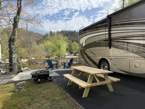 Creekside lot with class A motorhome showing picnic table, firepit and blue adirondack chairs by fence overlooking creek and resort